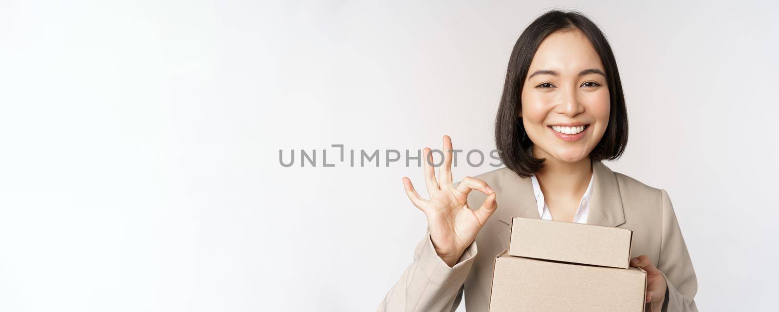Smiling asian businesswoman, showing okay sign and boxes with delivery goods, prepare order for client, standing over white background.