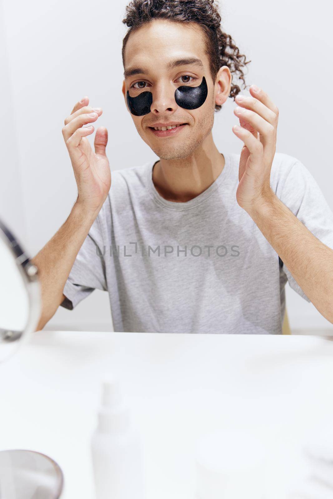 guy with curly hair in a white T-shirt sits at a table with a mirror and a towel face patches skin care by SHOTPRIME