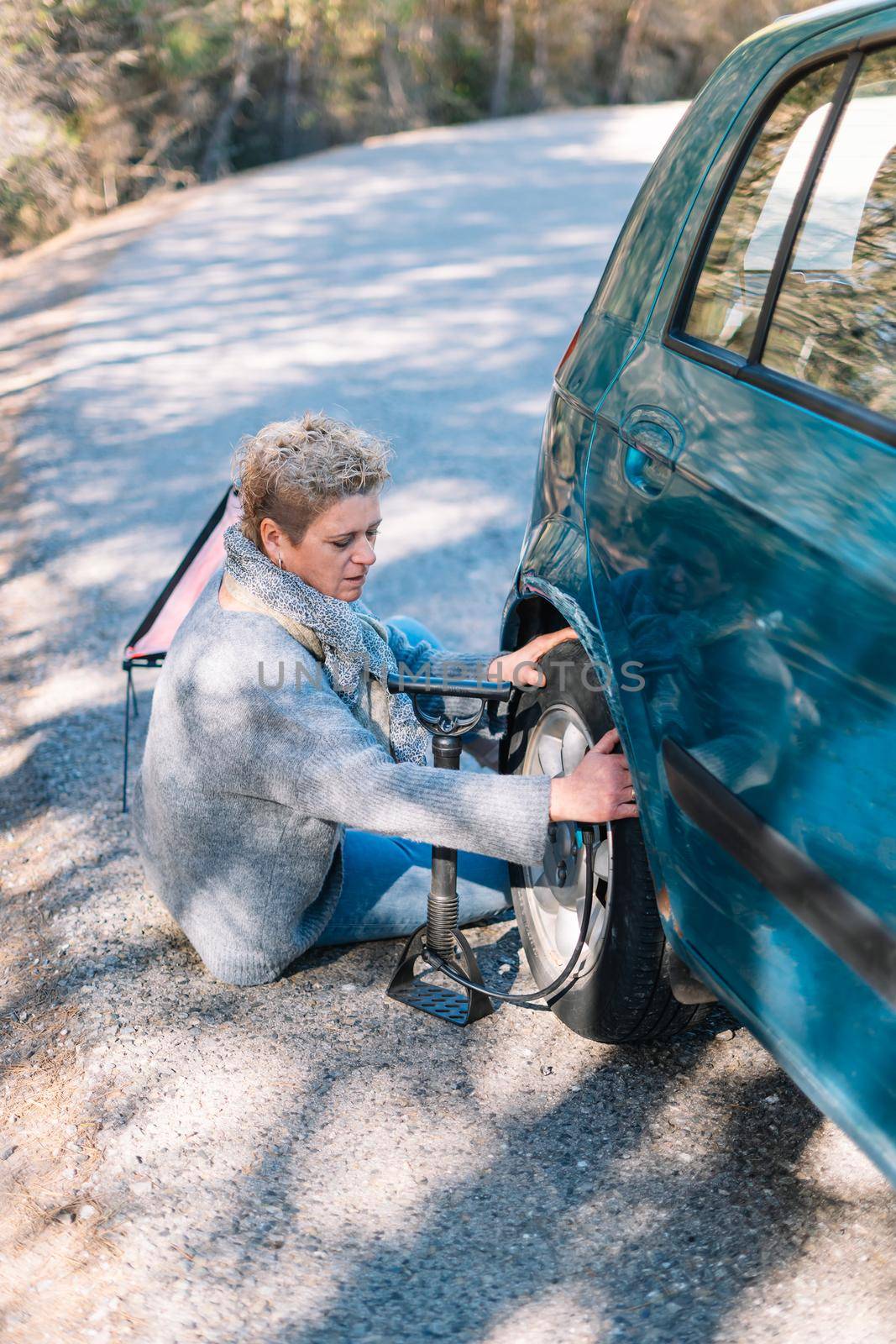 Adult blonde woman in blue jumper in foreground, inflating car tyre on the road after getting a puncture, on a sunny summer day with blue sky and natural light, with forest and mountain in the background.