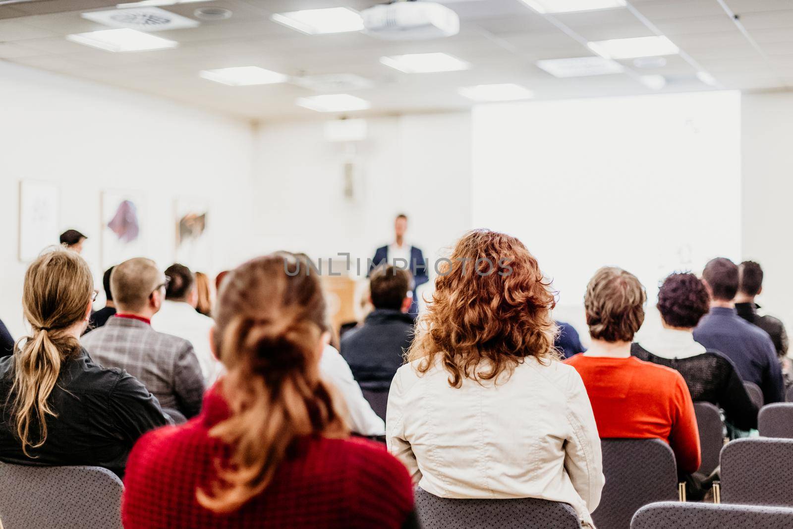 Business and entrepreneurship symposium. Speaker giving a talk at business meeting. Audience in conference hall. Rear view of unrecognized participant in audience.