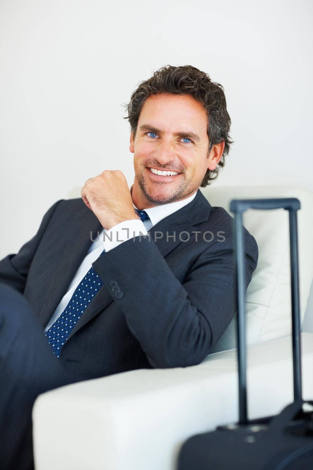 Smiling middle aged executive sitting on a chair with travel suitcase.