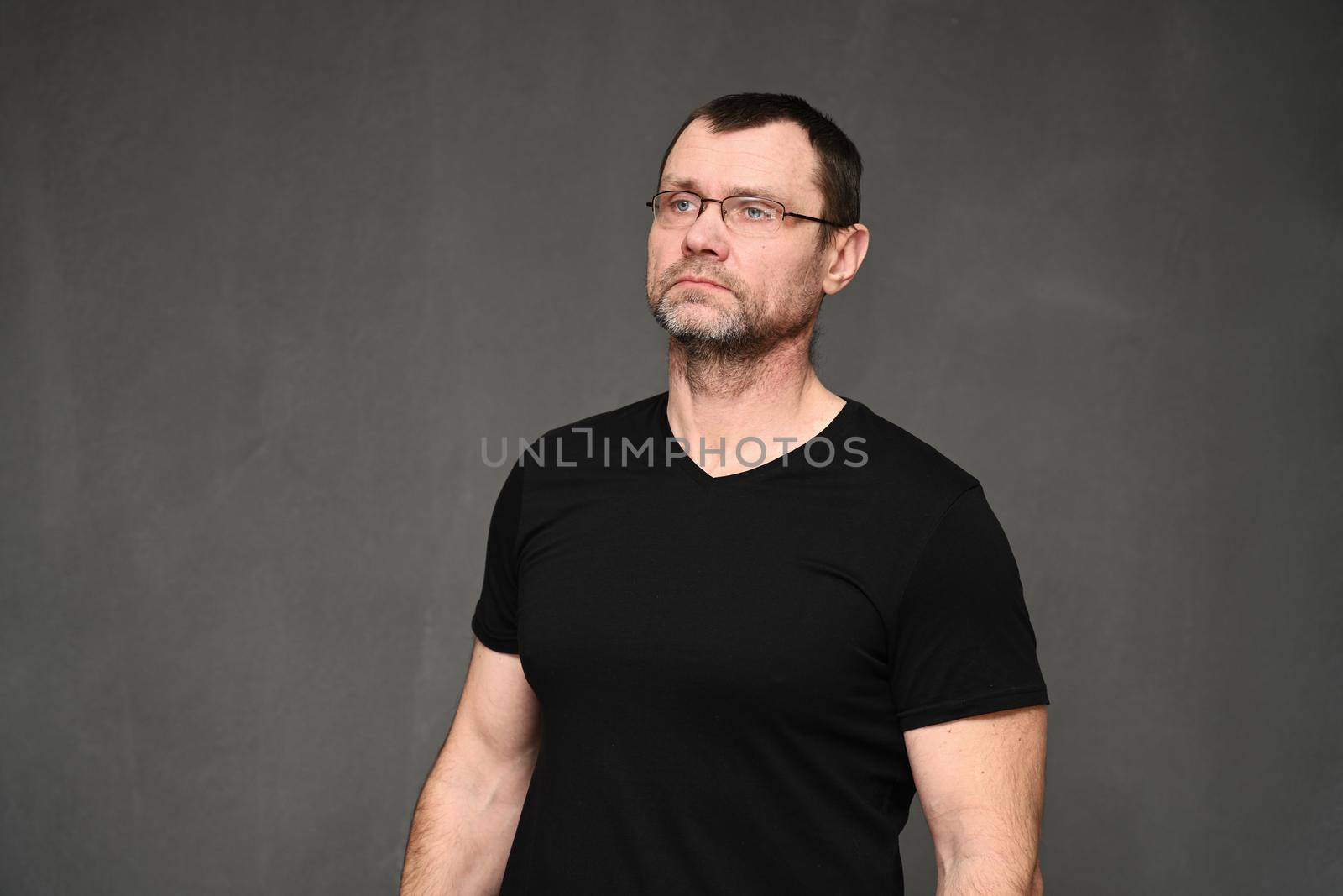 An adult man with glasses looks away. Portrait of a caucasian man in a black t-shirt on a gray background in the studio by chichaevstudio