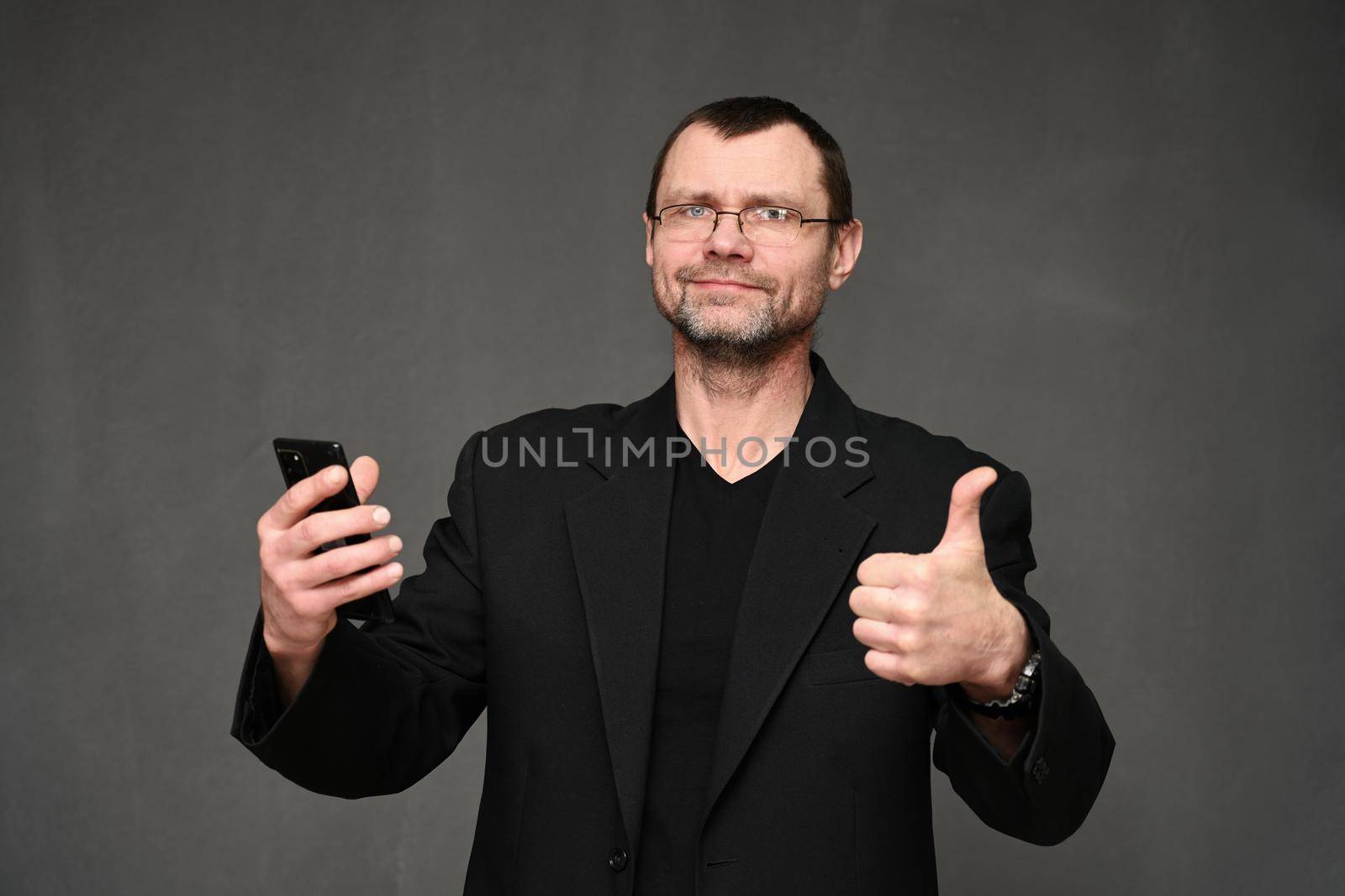 Caucasian businessman in a jacket looks at the camera with a smartphone in his hand and shows ok. Portrait on a gray background in the studio