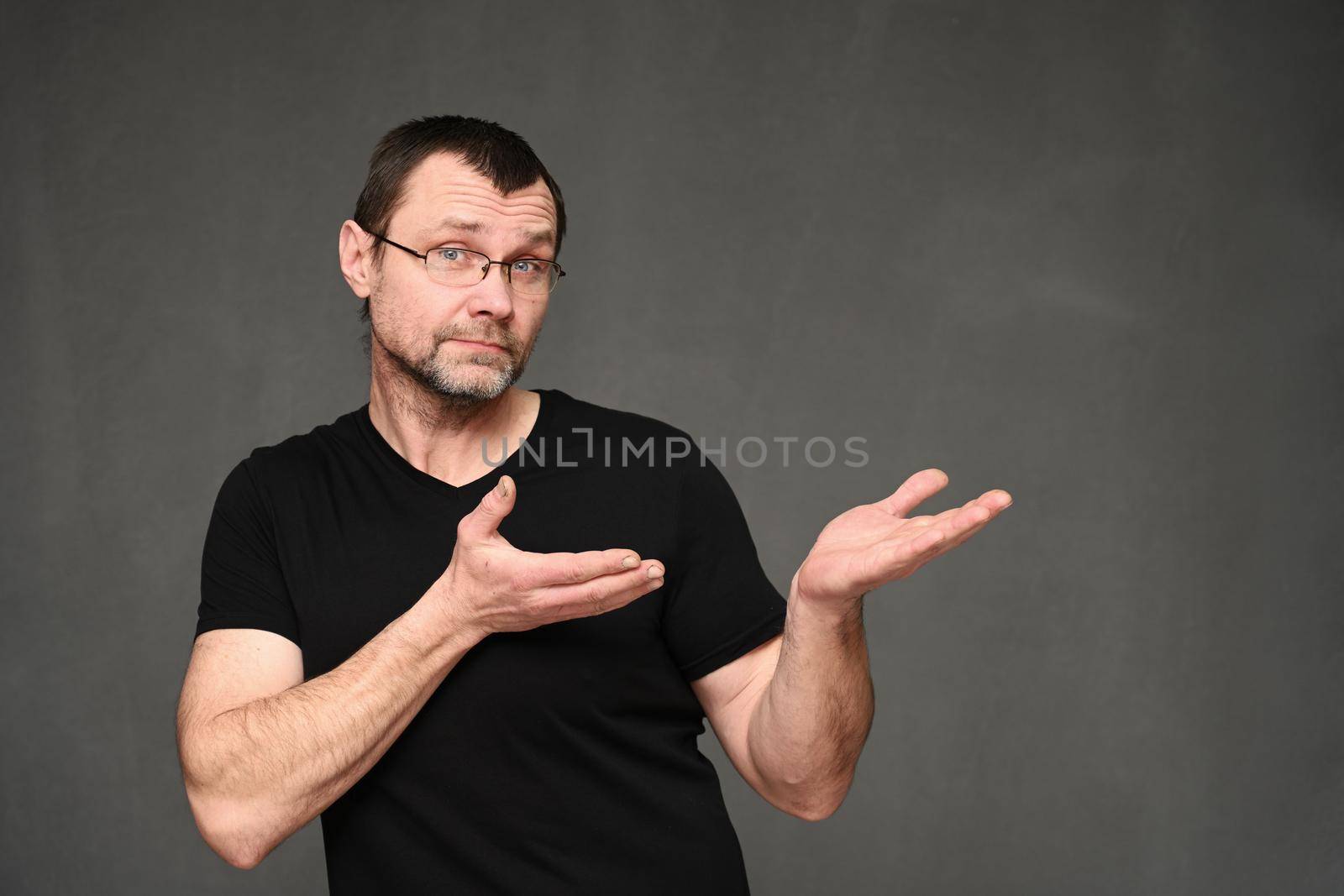 An adult man in glasses shows with emotions his hands to the side. Portrait of a caucasian man in a black t-shirt on a gray background in the studio