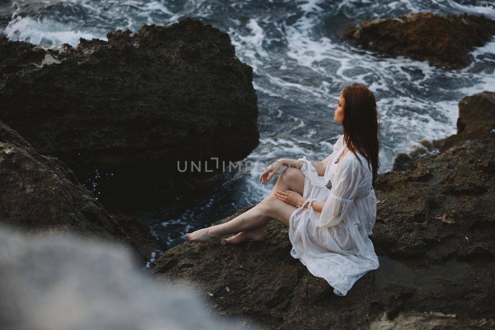 Barefoot Woman in White Dress with Wet Hair Sits on a Cliff. High quality photo