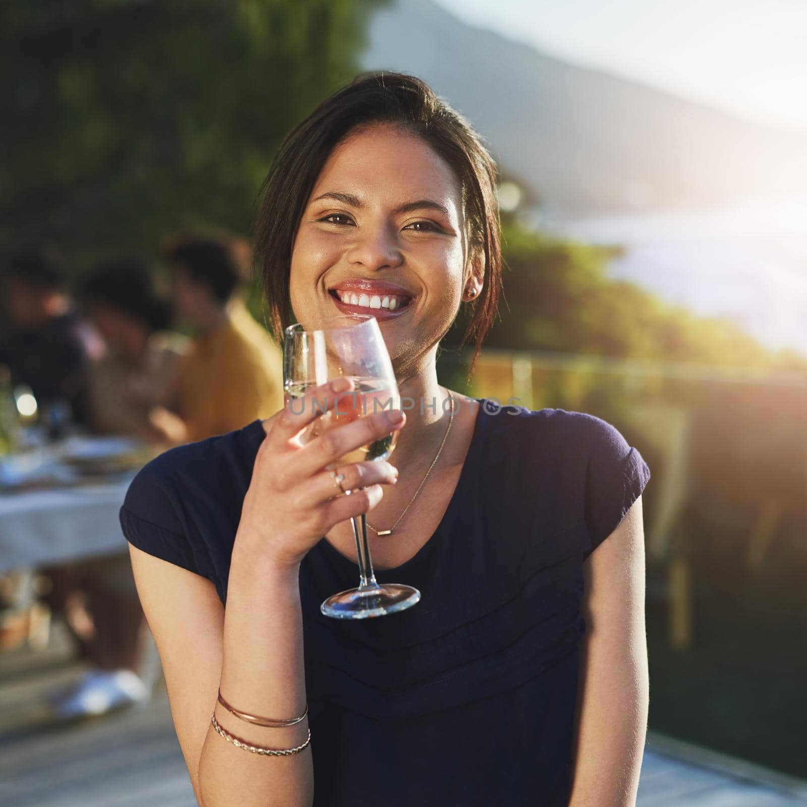 Shes as fine as the crisp wine she drinks. Shot of an attractive young woman enjoying a glass of wine outdoors with her friends in the background. by YuriArcurs