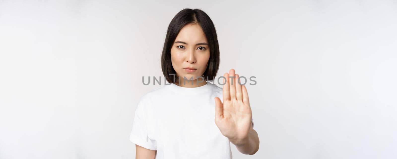 Image of asian girl showing stop, prohibit smth, extend one arm to show forbidding, taboo gesture, standing in tshirt over white background.