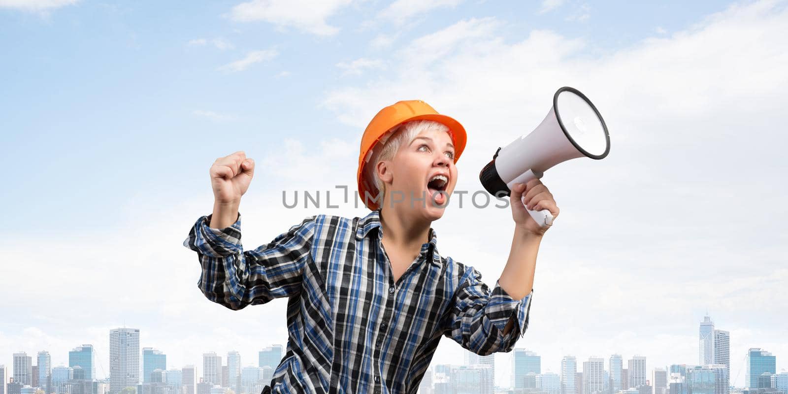 Expressive woman in safety helmet shouting into megaphone. Portrait of young emotional construction worker with wide open mouth on background of modern city. News announcement and advertisement.