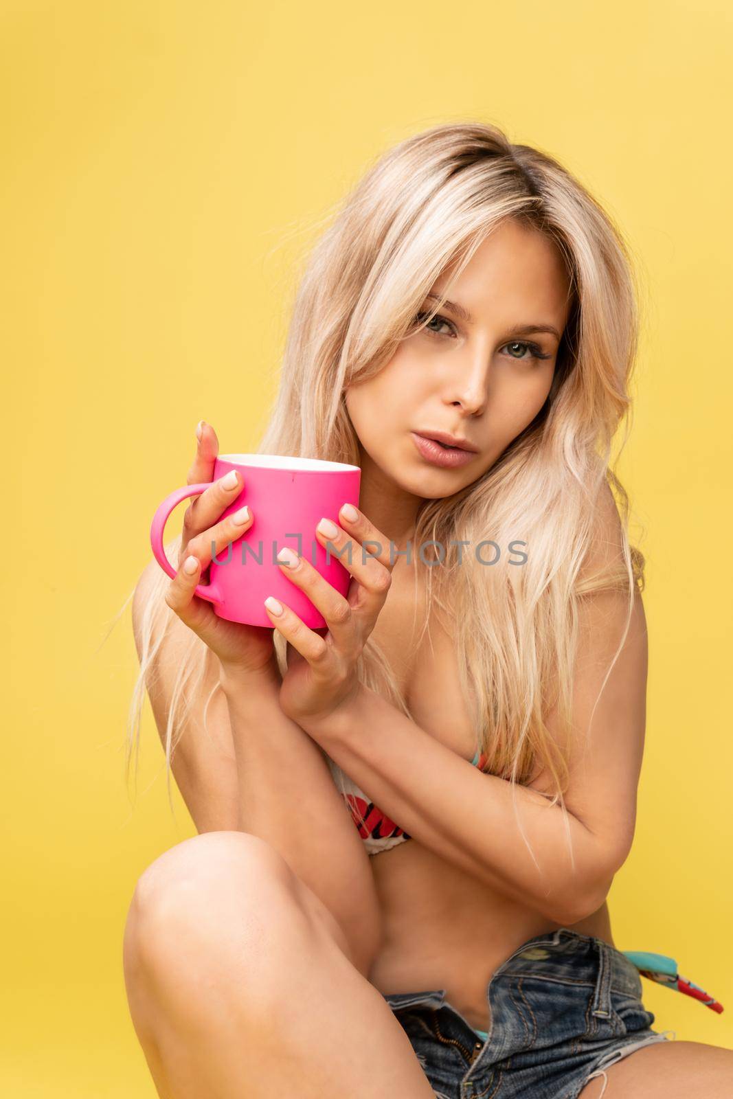 A girl sitting on the floor with her feet cross-legged keeps her head on a beautiful yellow background with a cup in hand yellow or pink and red playful swimsuit sexy, lady beautiful slim attractive white, lingerie. Vacation tan busty, cute soppy