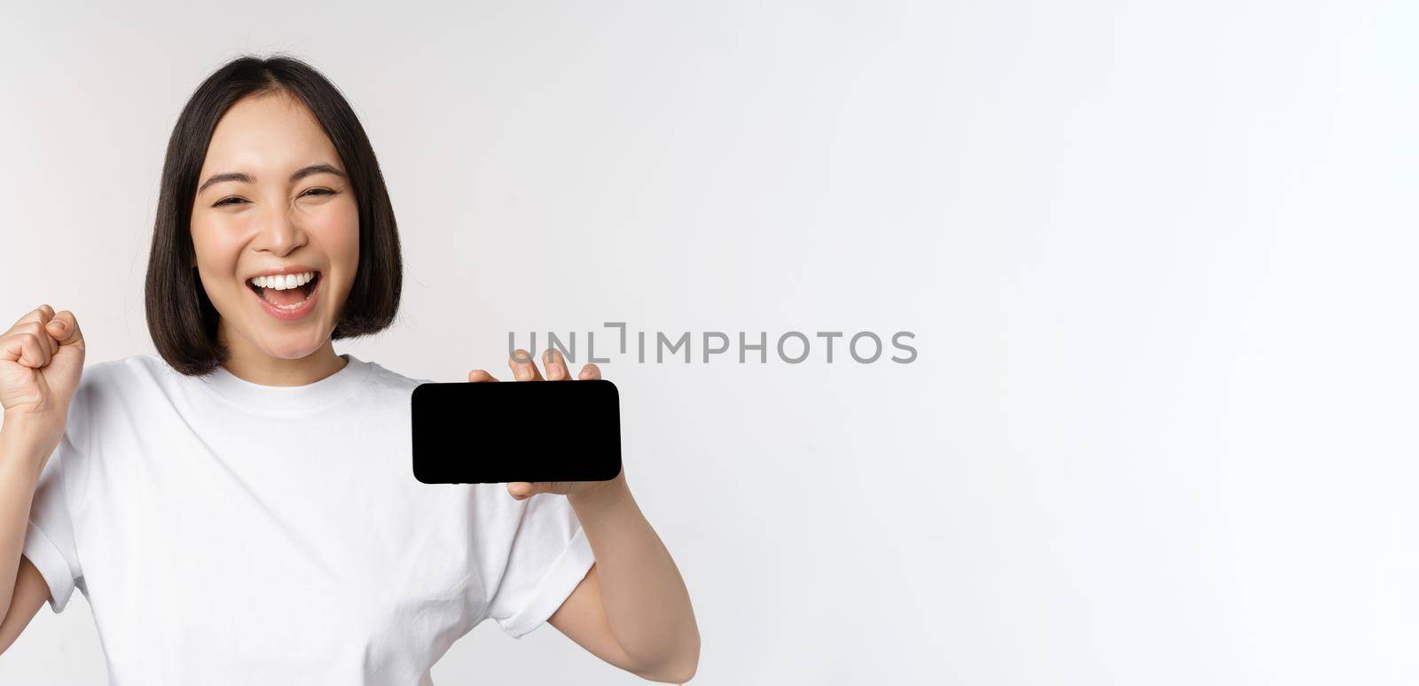 Enthusiastic asian girl scream in joy, showing horizontal smartphone screen, mobile phone display, standing over white background by Benzoix