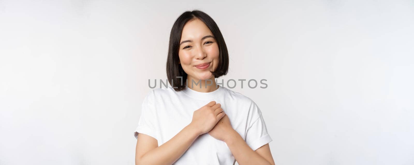 Portrait of carig young asian woman holding hands on heart, gazing with care and love, touched by something, feel flattered, standing over white background by Benzoix