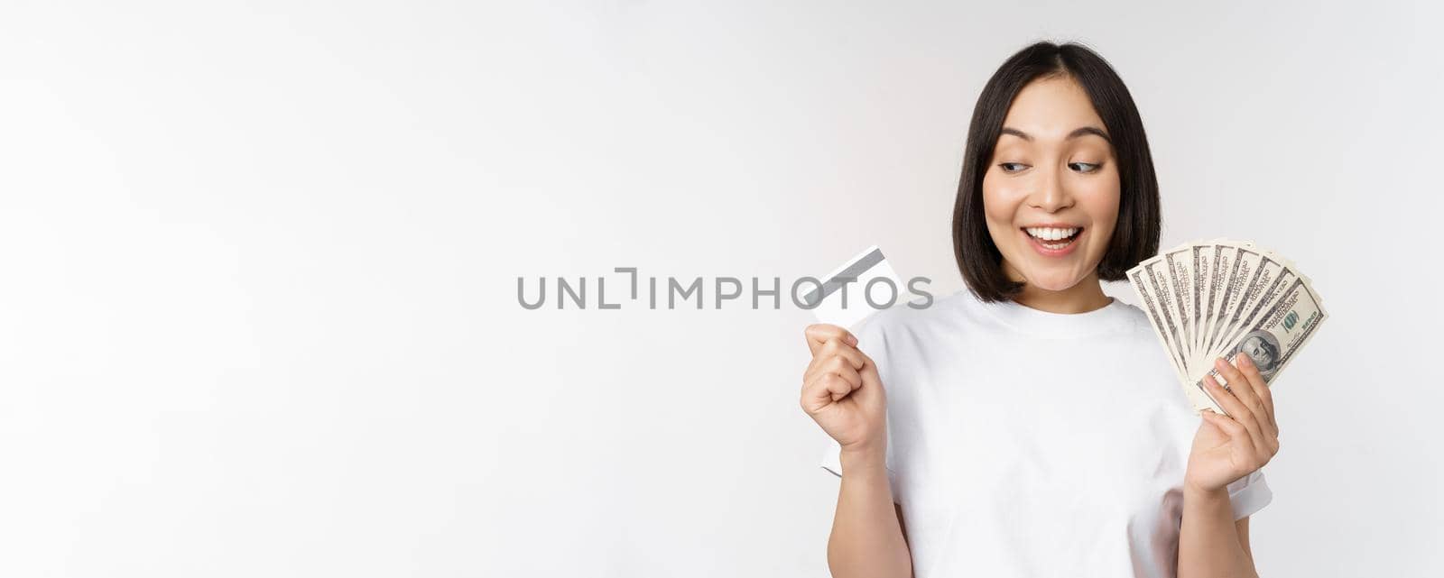 Portrait of asian woman smiling, holding credit card and money cash, dollars, standing in tshirt over white background by Benzoix