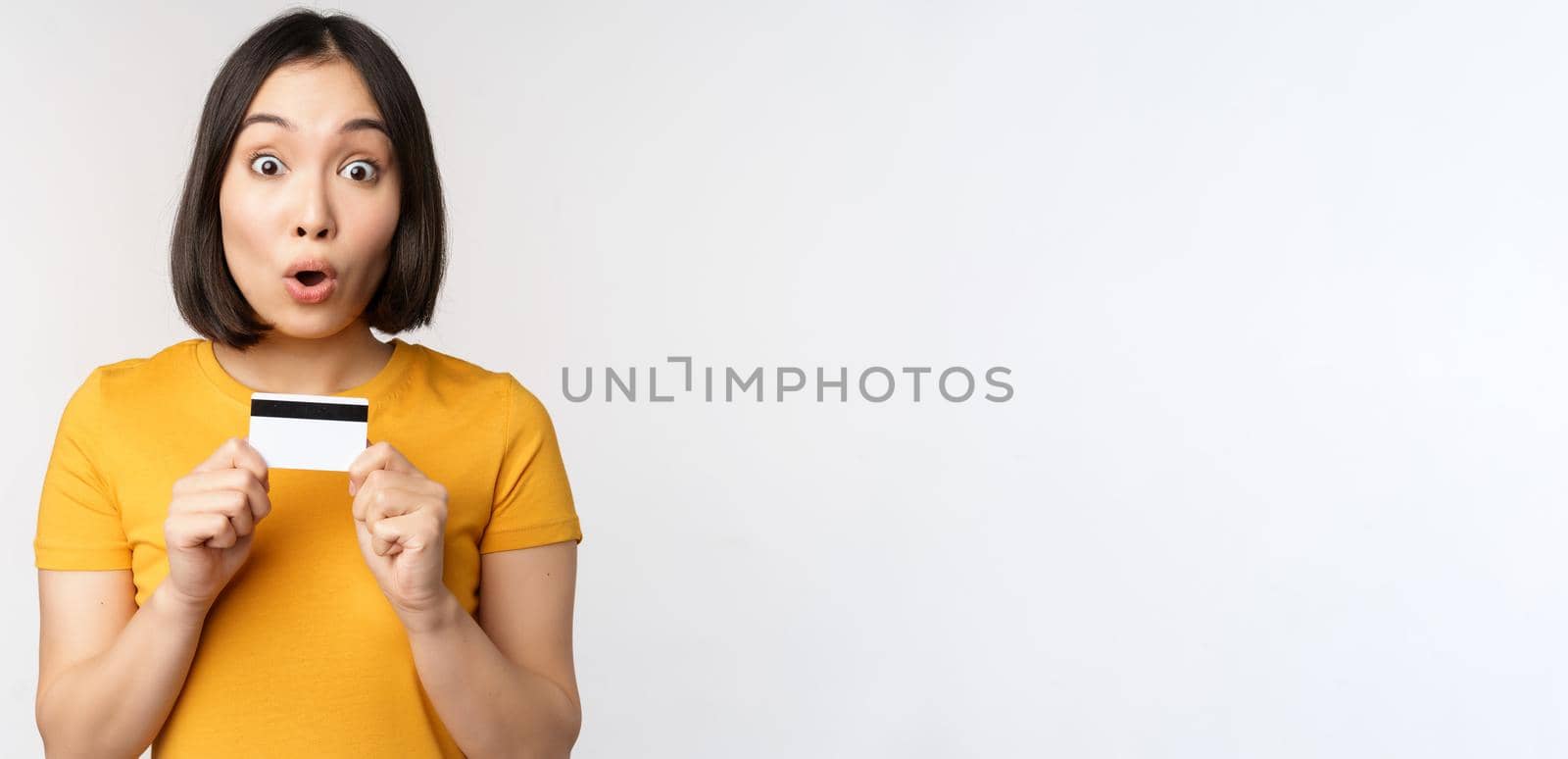 Portrait of beautiful korean girl holding credit card, recommending bank service, standing in yellow tshirt over white background.