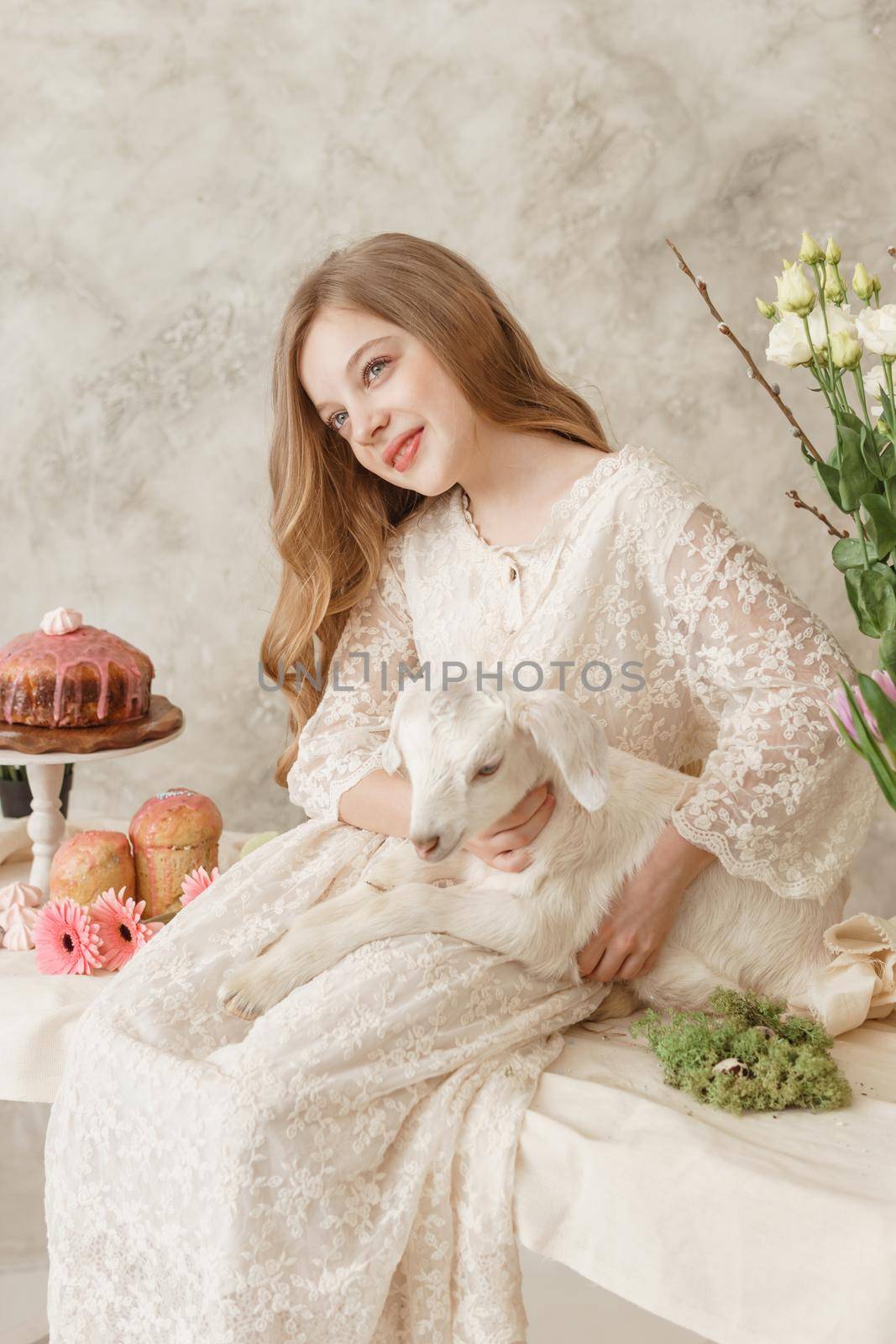 The girl is sitting at the Easter table with cakes, spring flowers and quail eggs and holding a white kid in her arms. Happy Easter celebration