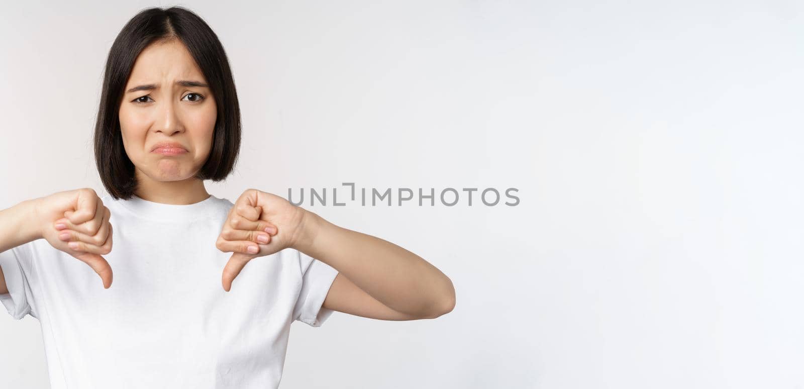 Image of asian woman showing thumbs down, dislike smth, looking disappointed, standing over white background. Copy space