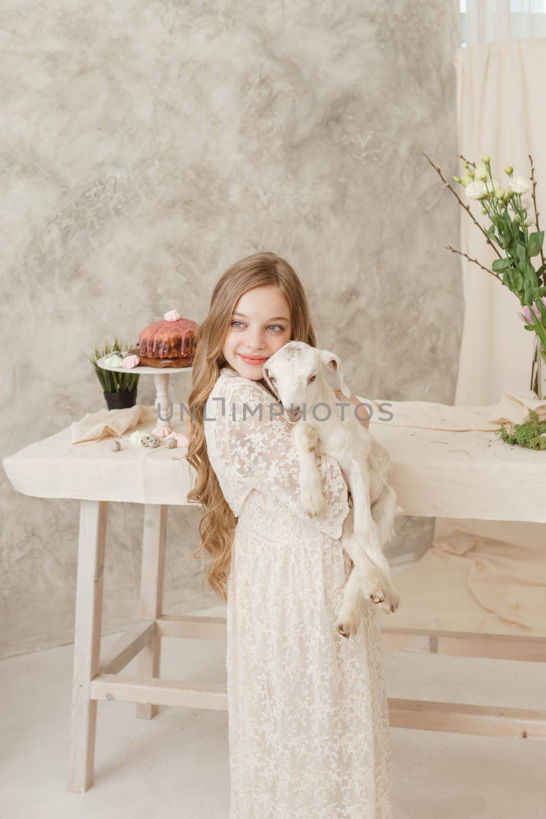 The girl is sitting at the Easter table with cakes, spring flowers and quail eggs and holding a white kid in her arms. Happy Easter celebration