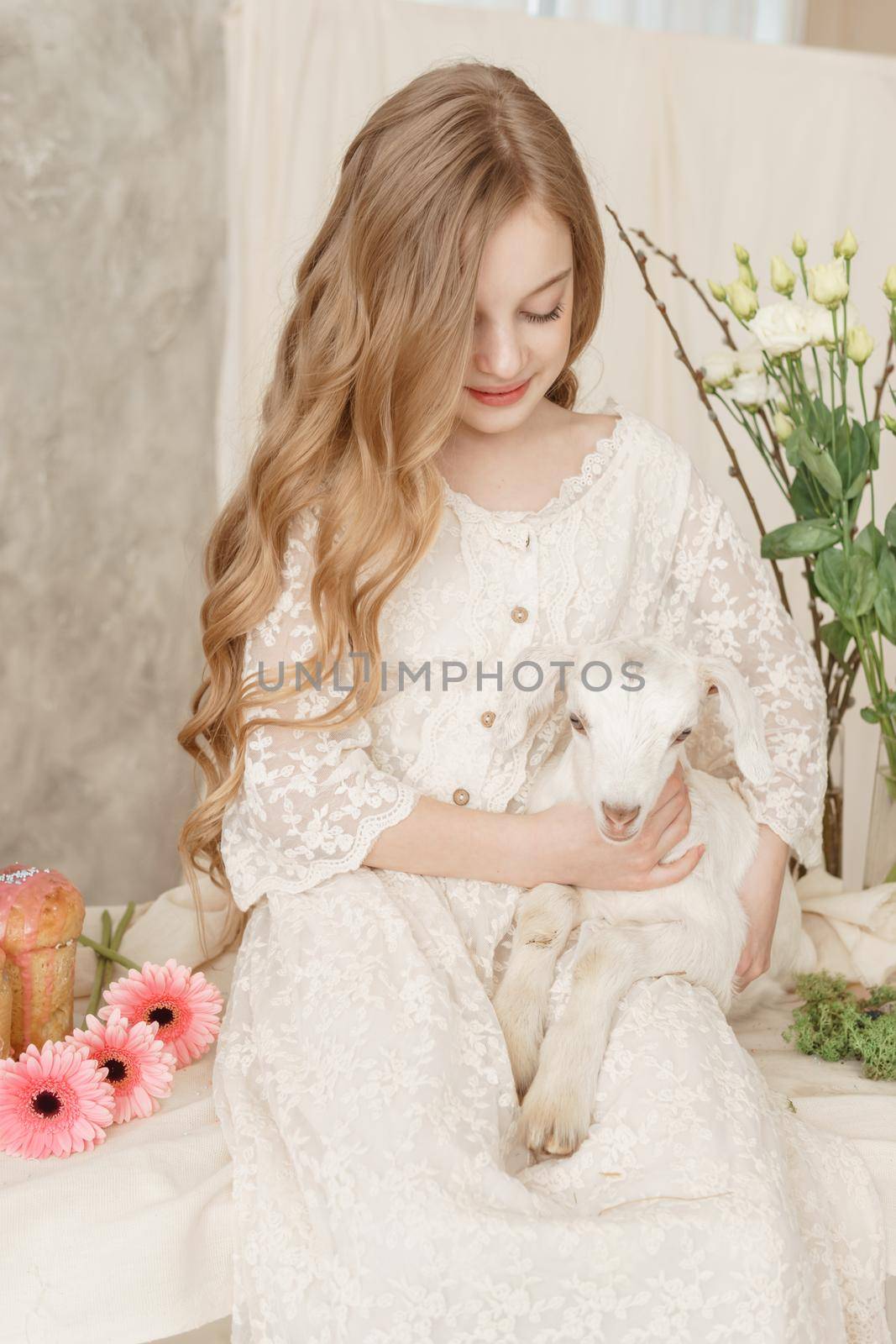 The girl is sitting at the Easter table with cakes, spring flowers and quail eggs and holding a white kid in her arms. Happy Easter celebration