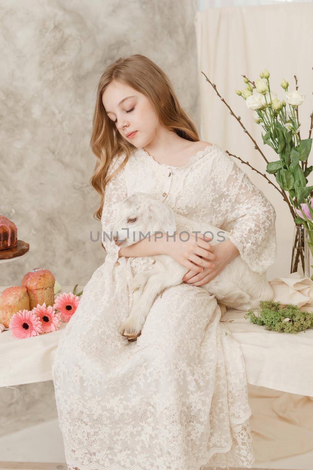 The girl is sitting at the Easter table with cakes, spring flowers and quail eggs and holding a white kid in her arms. Happy Easter celebration