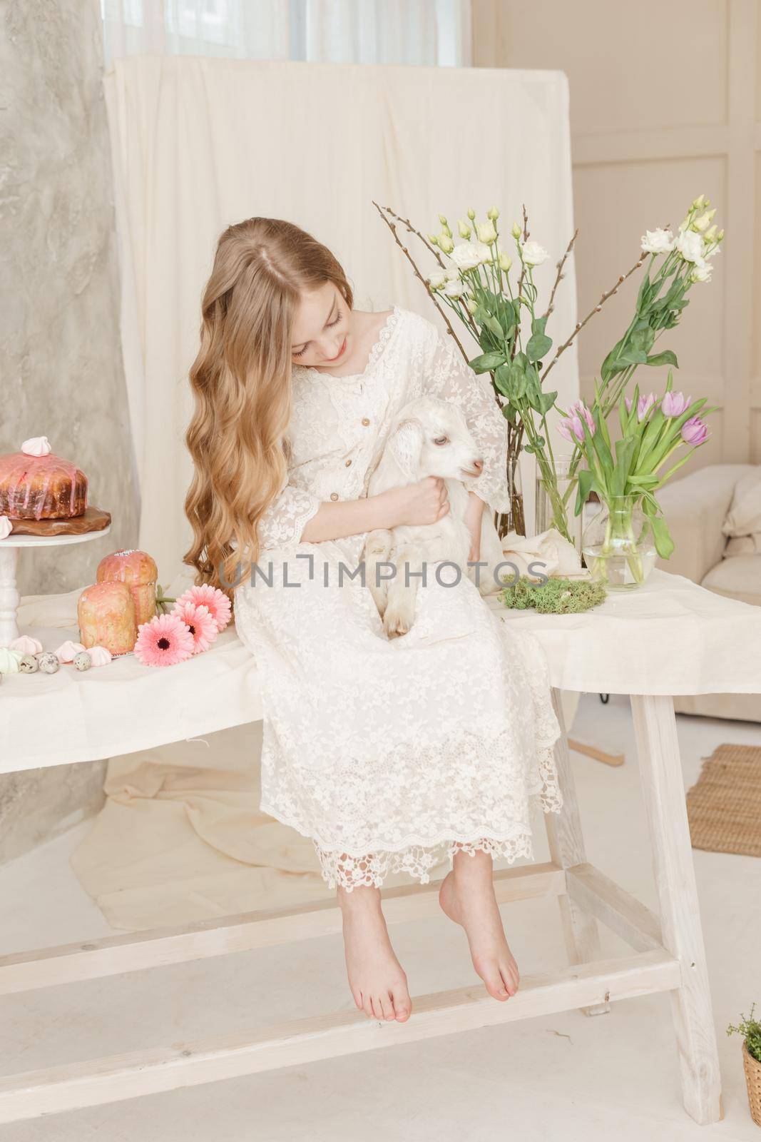 The girl is sitting at the Easter table with cakes, spring flowers and quail eggs and holding a white kid in her arms. Happy Easter celebration