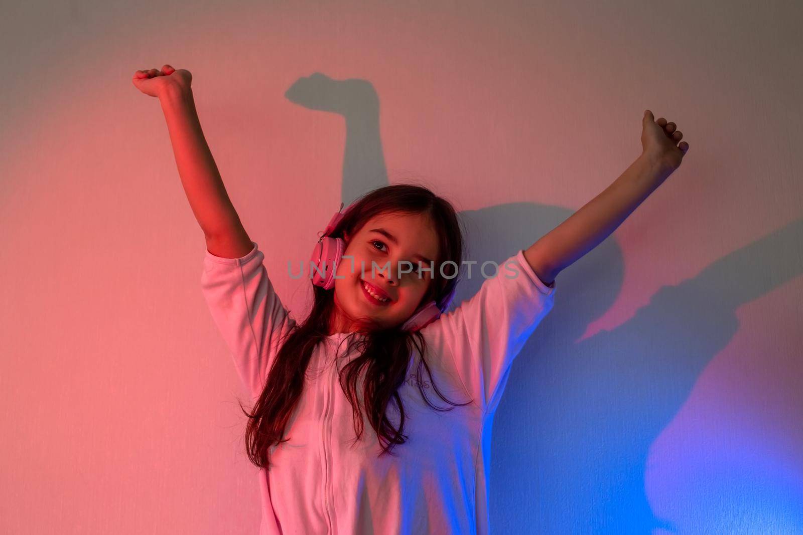Dancing happy little girl in pink headphones with loose dark hair in neon pink and blue light against the wall, raised her hands up