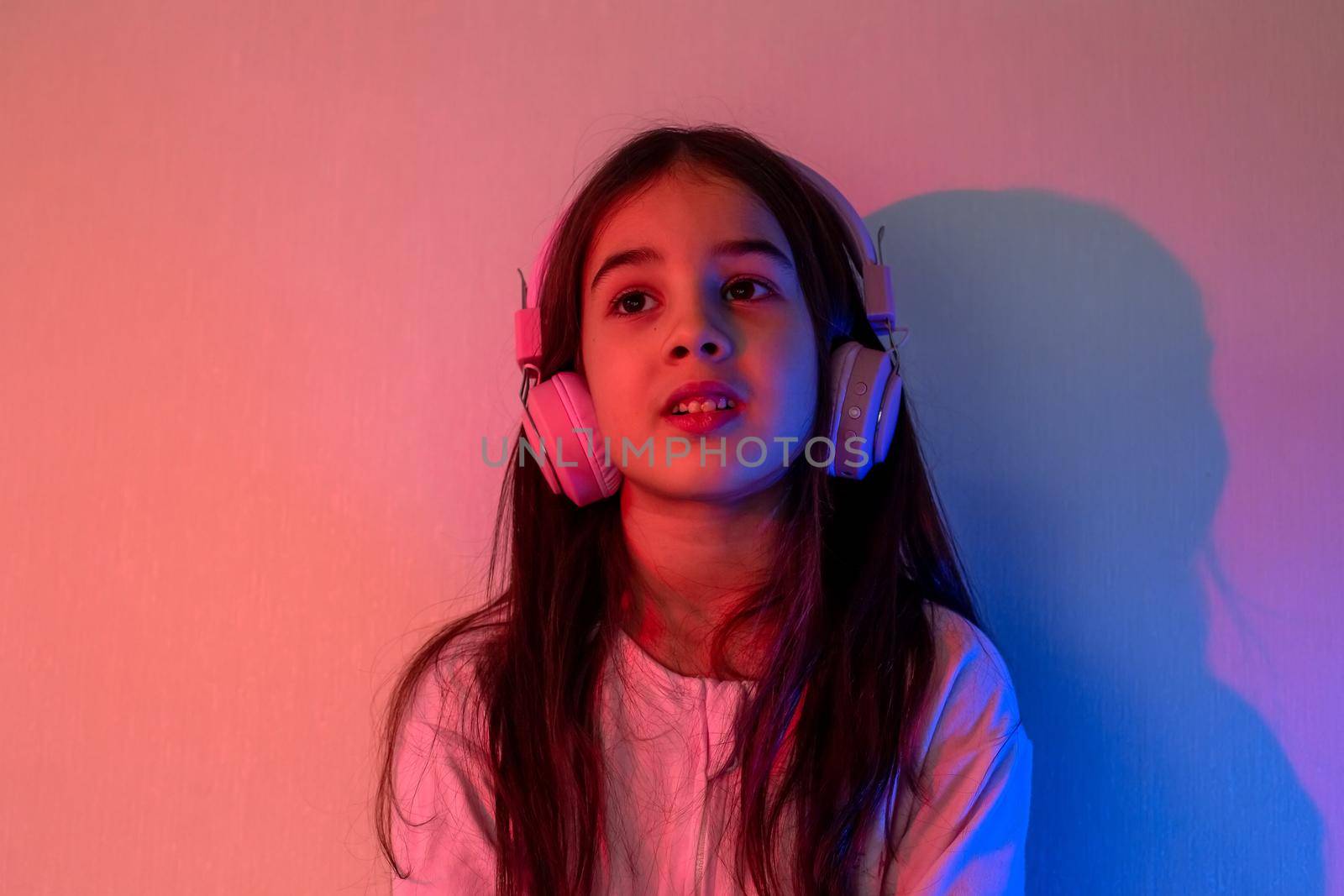 Portrait of a cute little girl in pink headphones, with loose dark hair, listening to music in neon pink blue light
