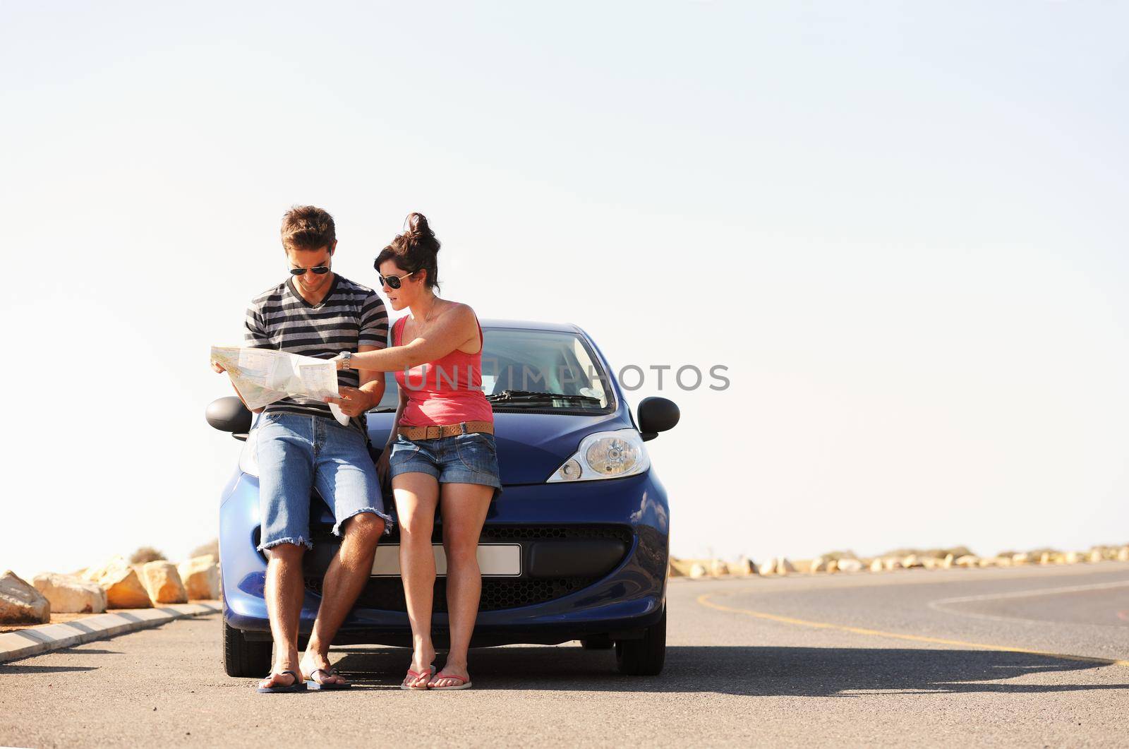 A man and a woman parked on the side of the road and reading a map together while seated on the bonnet of their car.