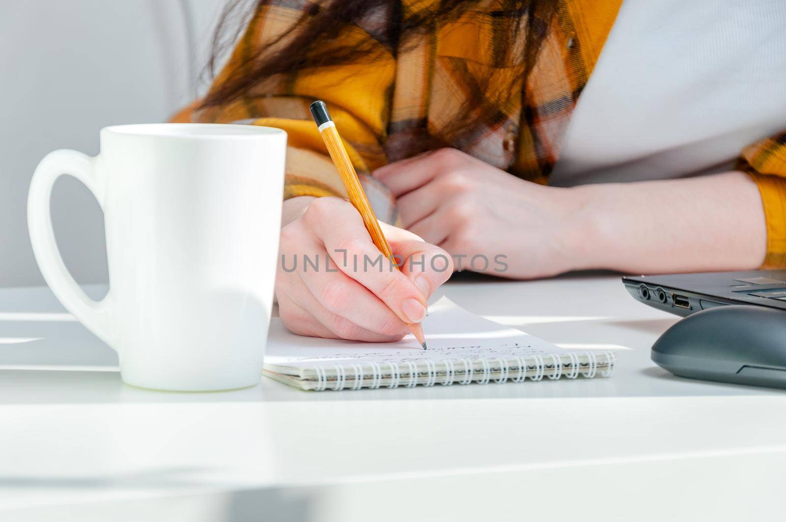 Close-up woman holding pencil in hand writing in notepad on workplace with coffee cup and laptop. Woman working at home.