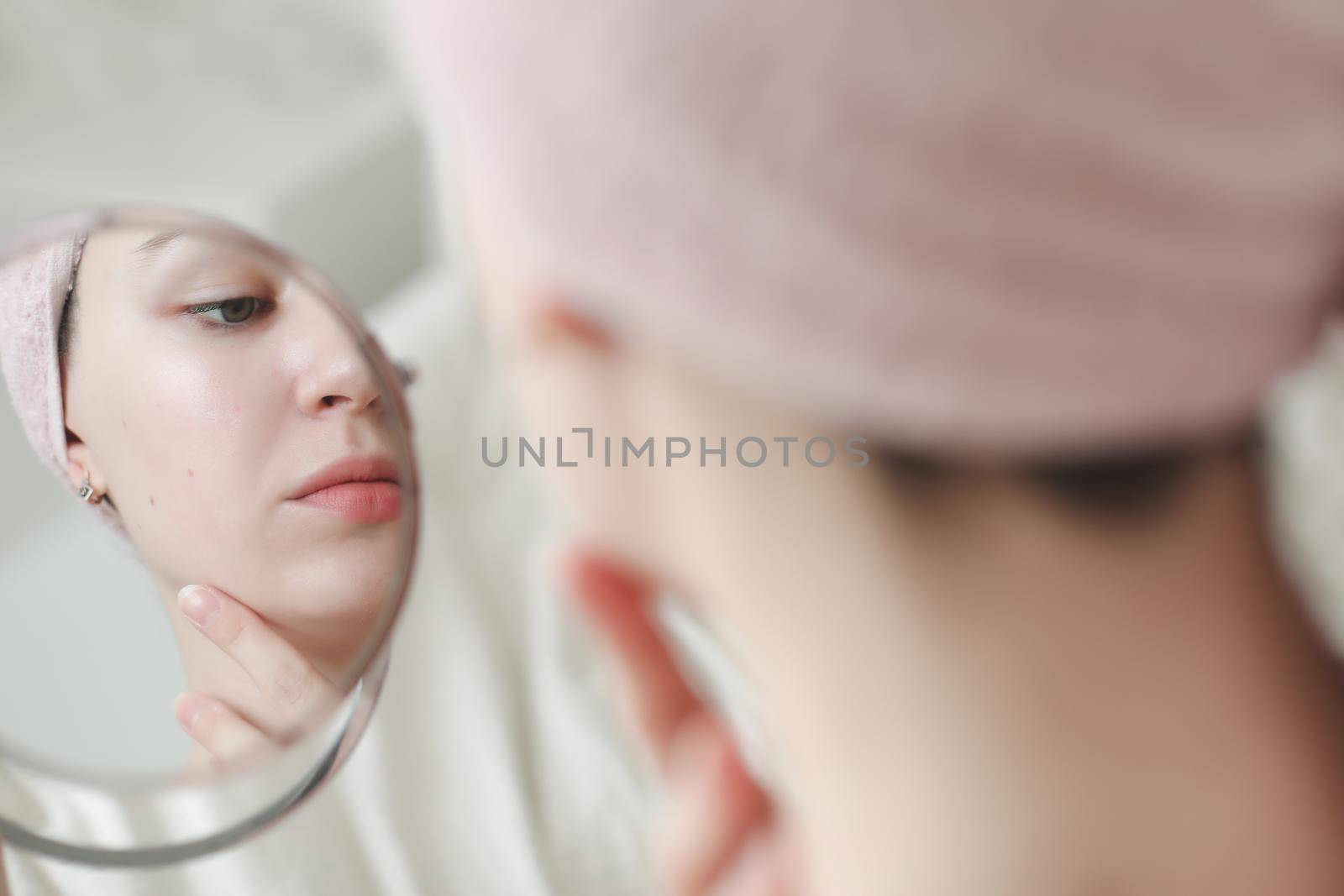 young woman with a towel on head looking in the mirror and apply cream on face skin.