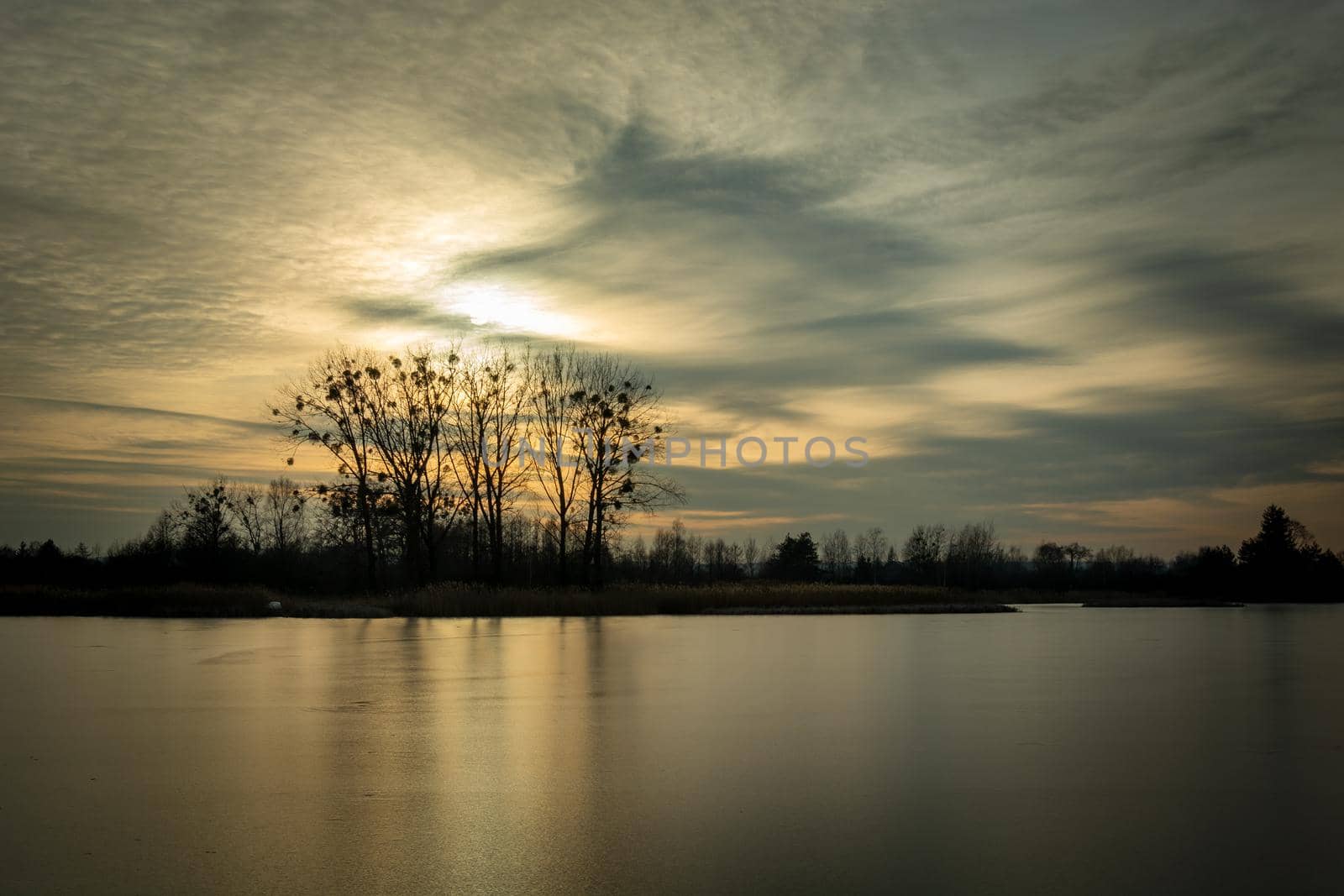 The sun behind the clouds over the frozen lake and trees on the shore by darekb22