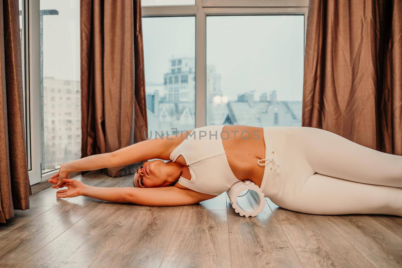 Adult athletic woman, in white bodysuit, performing fascia exercises on the floor - caucasian woman using a massage foam roller - a tool to relieve tension in the back and relieve muscle pain - the concept of physiotherapy and stretching training.