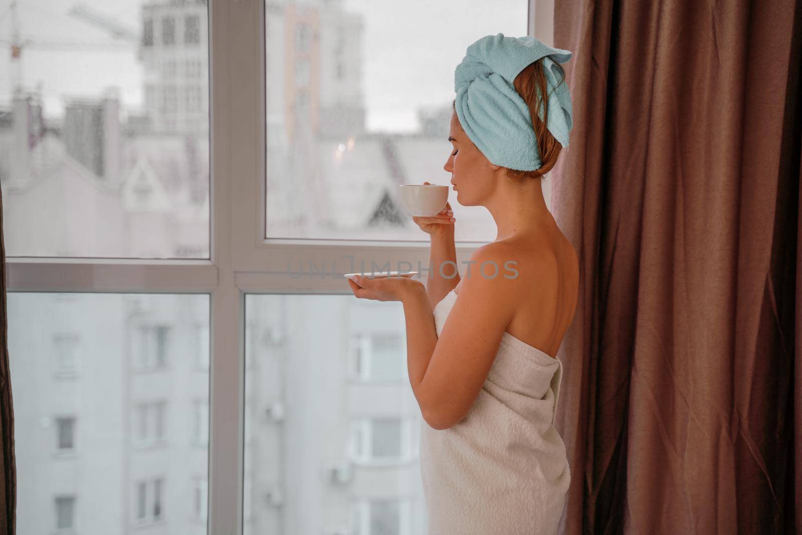 Young serene relaxed woman in spa bath towel drinking hot beverage tea coffee after taking shower bath at home. Beauty treatment, hydration concept. by Matiunina