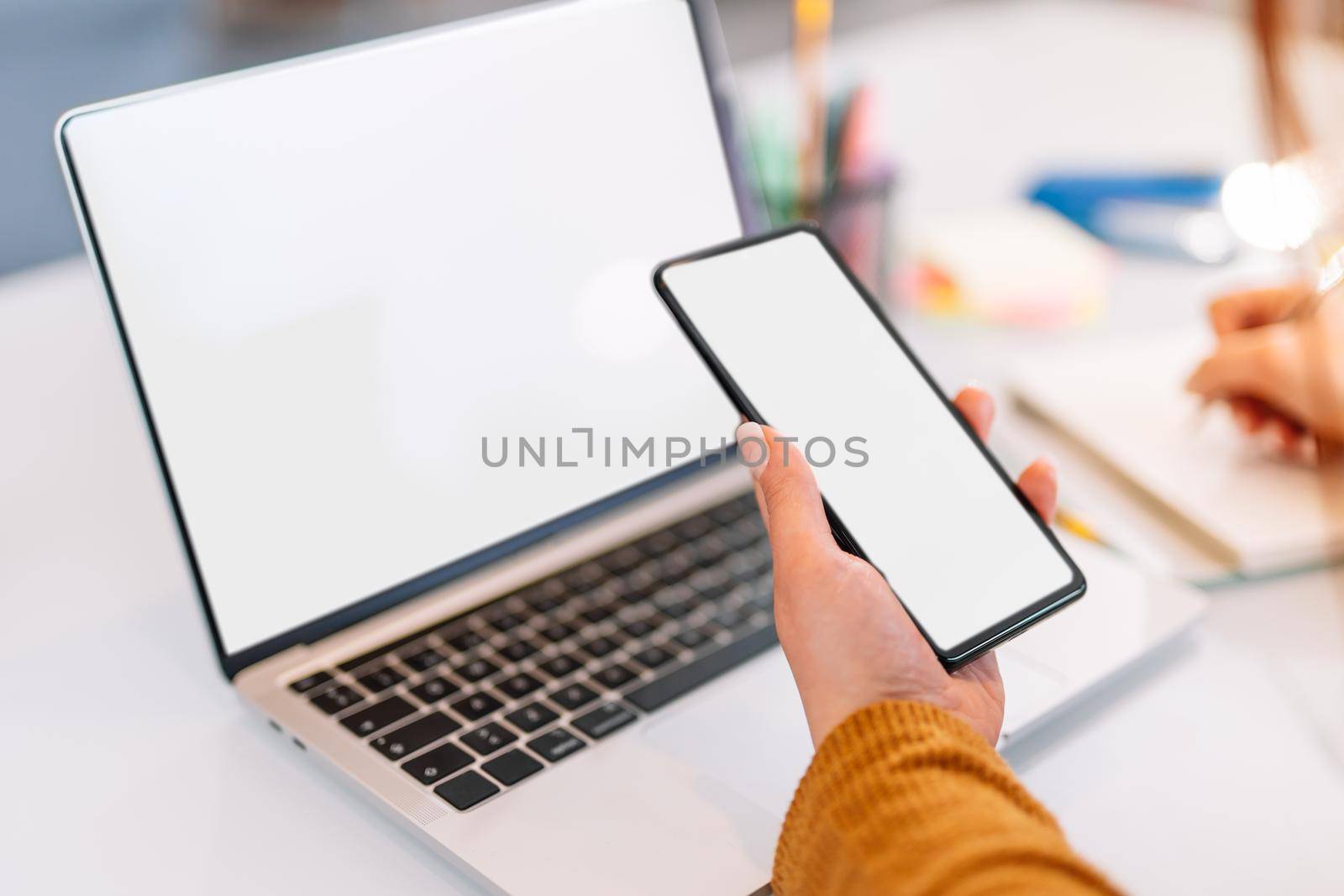 Hand shot of a young girl writing work matters in a notebook while consulting her laptop computer and mobile phone. Large white table with a white laptop, post-its of various colours yellow, pink and a jar with various pens and markers. Natural light illumination. Young woman with yellow sweater.