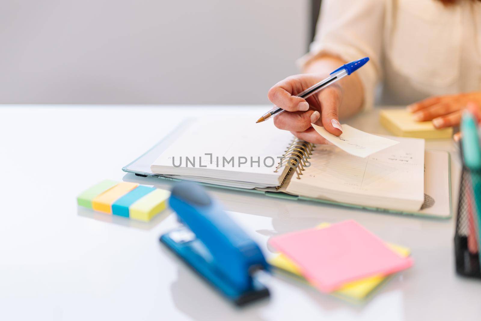 Hand shot of a young girl writing work matters in a notebook while consulting her laptop computer. Large white table with a white laptop, post-its of various colours yellow, pink and a jar with various pens and markers. Natural light illumination