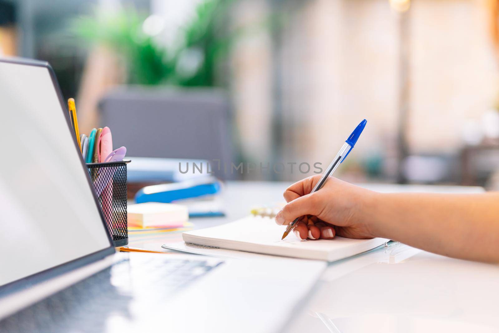 Hand shot of a young girl writing work matters in a notebook while consulting her laptop computer. Large white table with a white laptop, post-its of various colours yellow, pink and a jar with various pens and markers. Natural light illumination