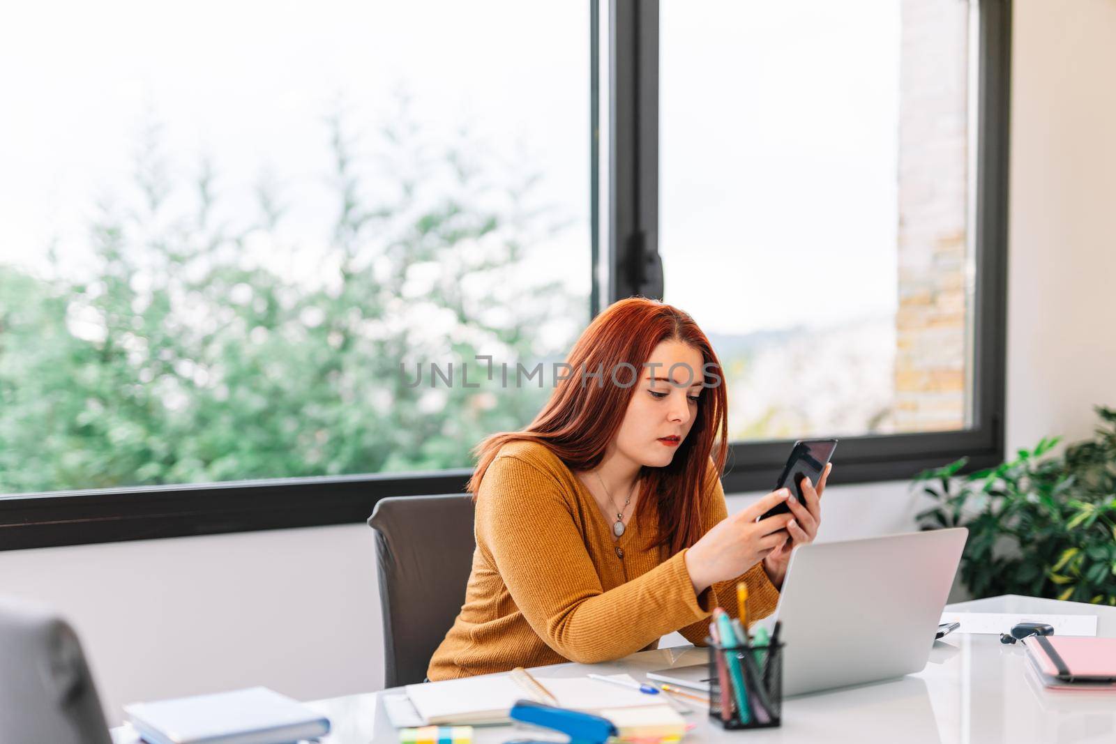 Pretty young red-haired girl in yellow sweater chatting on her smartphone while working on her laptop. Young businesswoman teleworking from home in a very bright environment with a large window in the background and natural light.