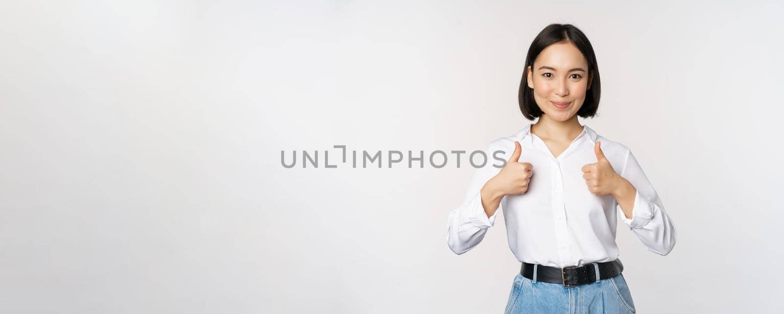Image of beautiful adult asian woman showing thumbs up, wearing formal office, university clothing, recommending company, standing over white background by Benzoix