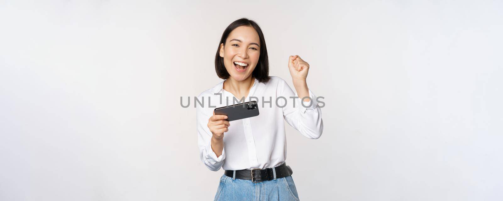 Happy asian woman do winner dance, triumphing, winning on mobile phone video game, holding smartphone horizontal position and celebrating, white background.