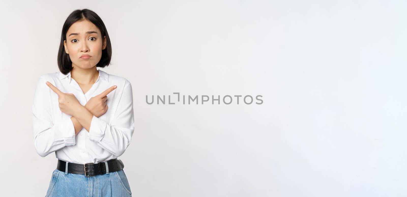 Confused sad asian girl cant decide, pointing fingers left and right, sulking and shrugging indecisive, cannot choose, standing over white background by Benzoix