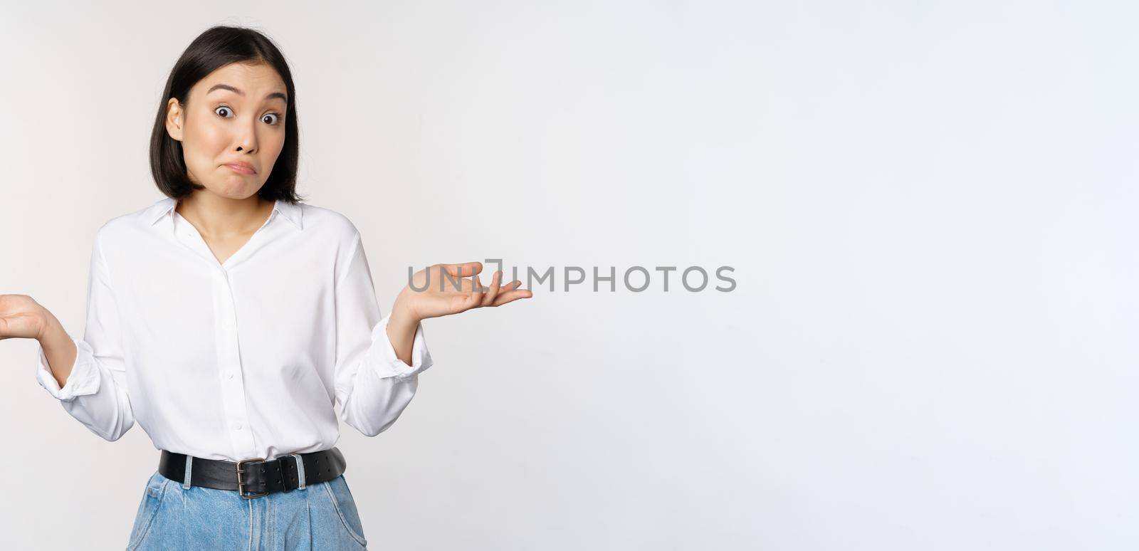 Image of confused asian office manager, office lady shrugging shoulders and looking clueless, standing puzzled against white background by Benzoix