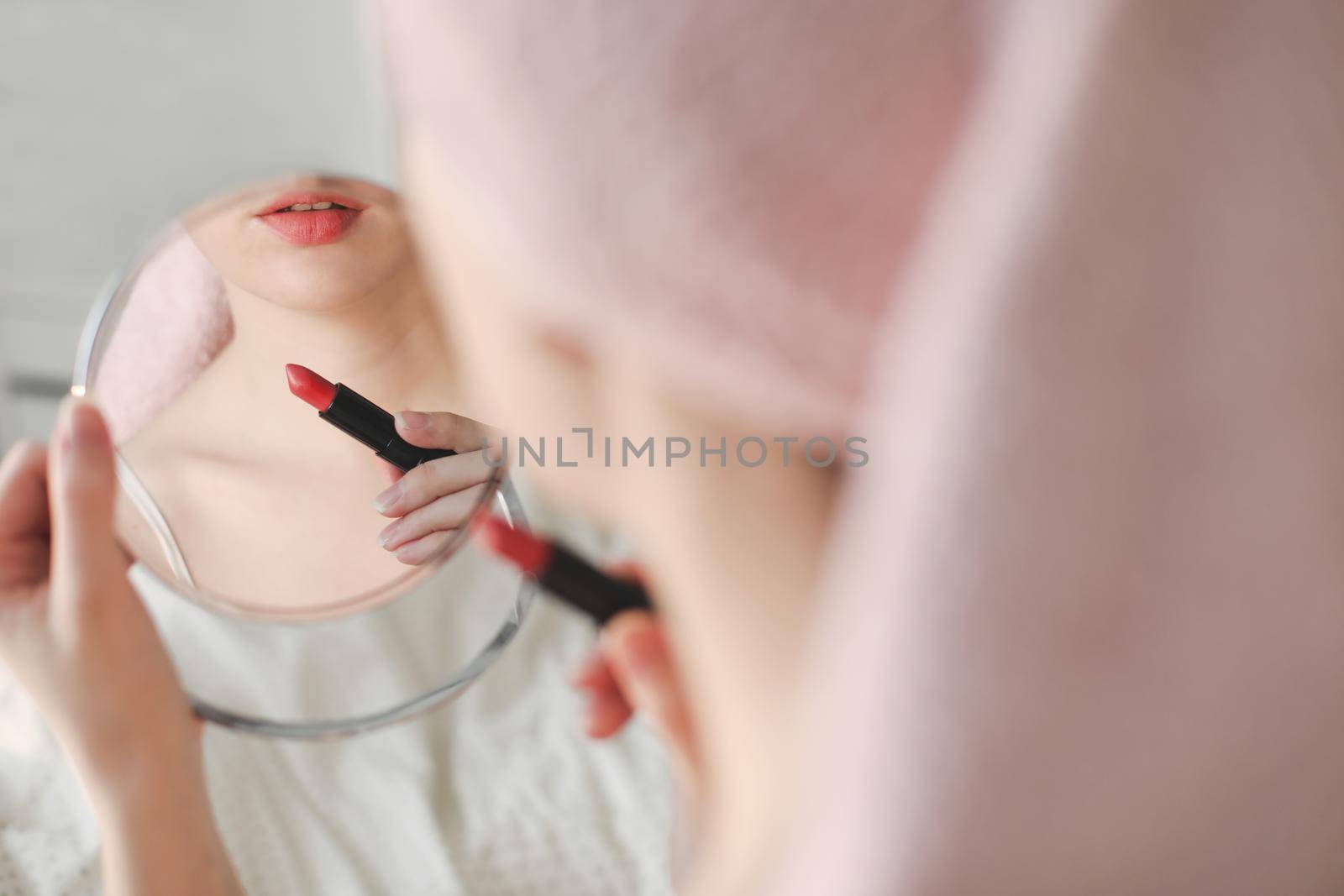 Beautiful attractive happy smiling woman paints lips with red lipstick using a small round mirror during home makeup in the morning.