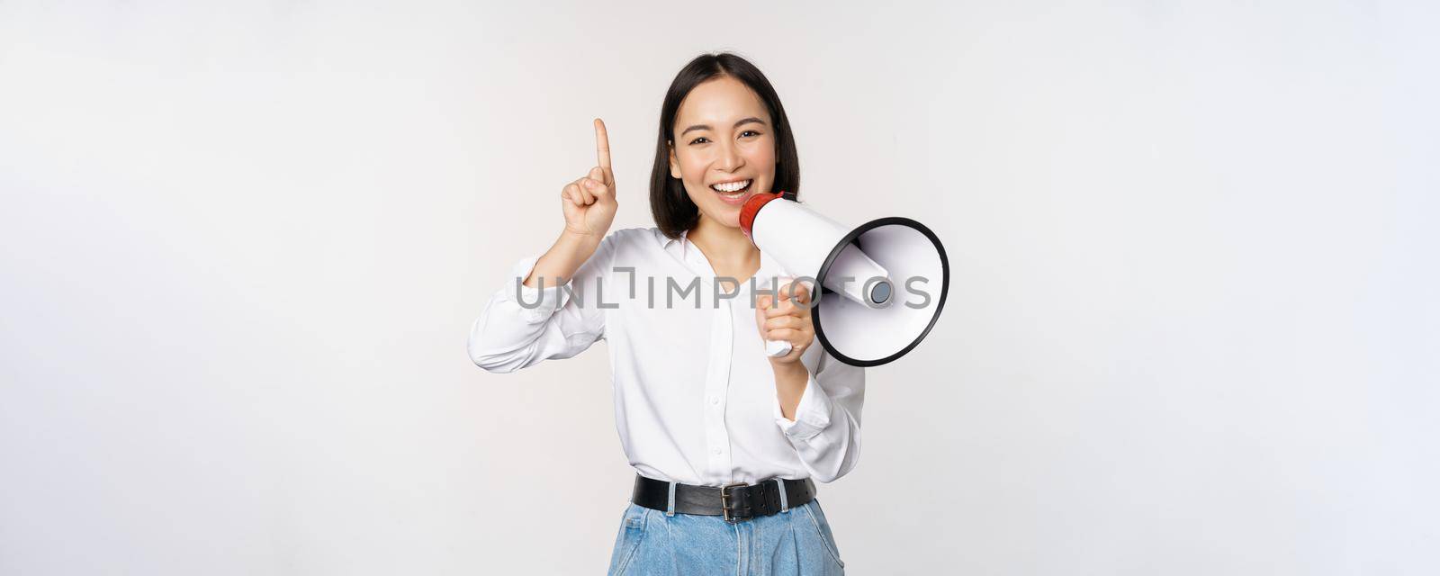Smiling happy asian girl talking in megaphone and pointing up, announcing discount promo, showing advertisement on top, standing over white background.