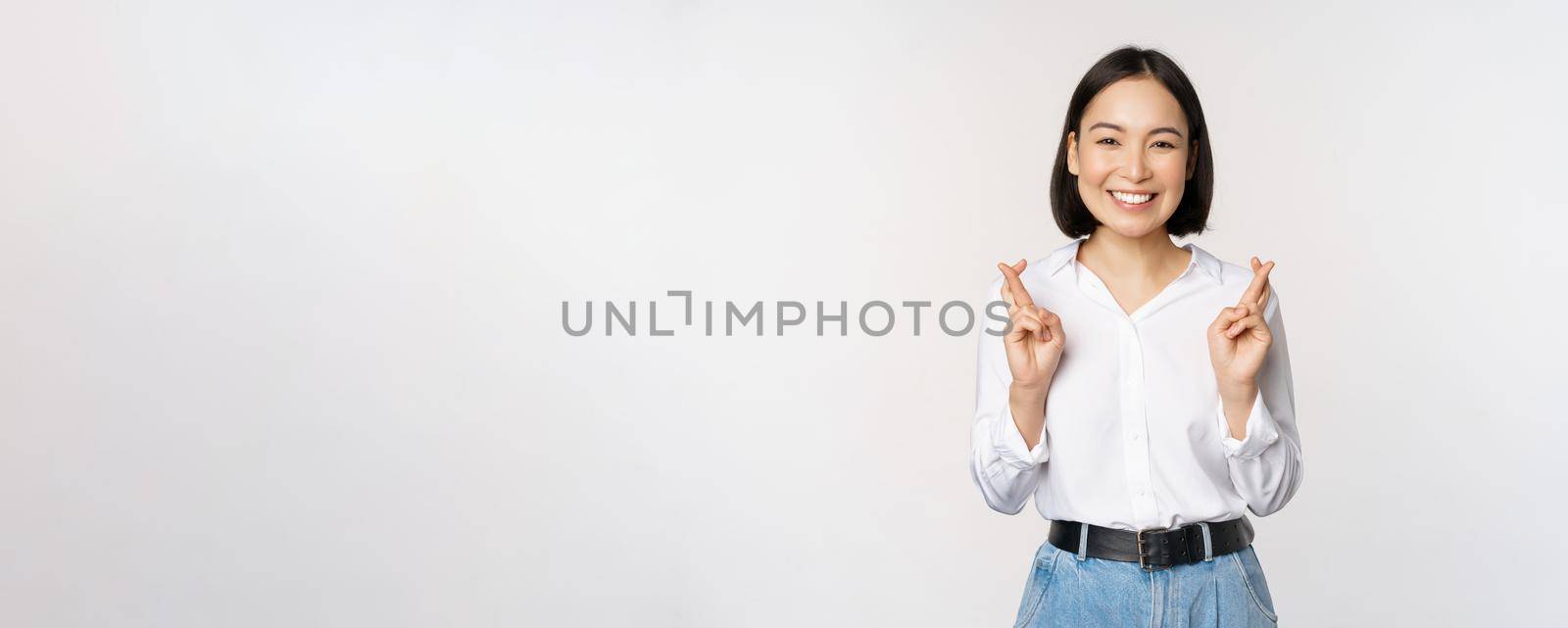 Portrait of young korean woman, asian girl cross fingers and praying, making wish, anticipating, waiting for results, standing over white background by Benzoix