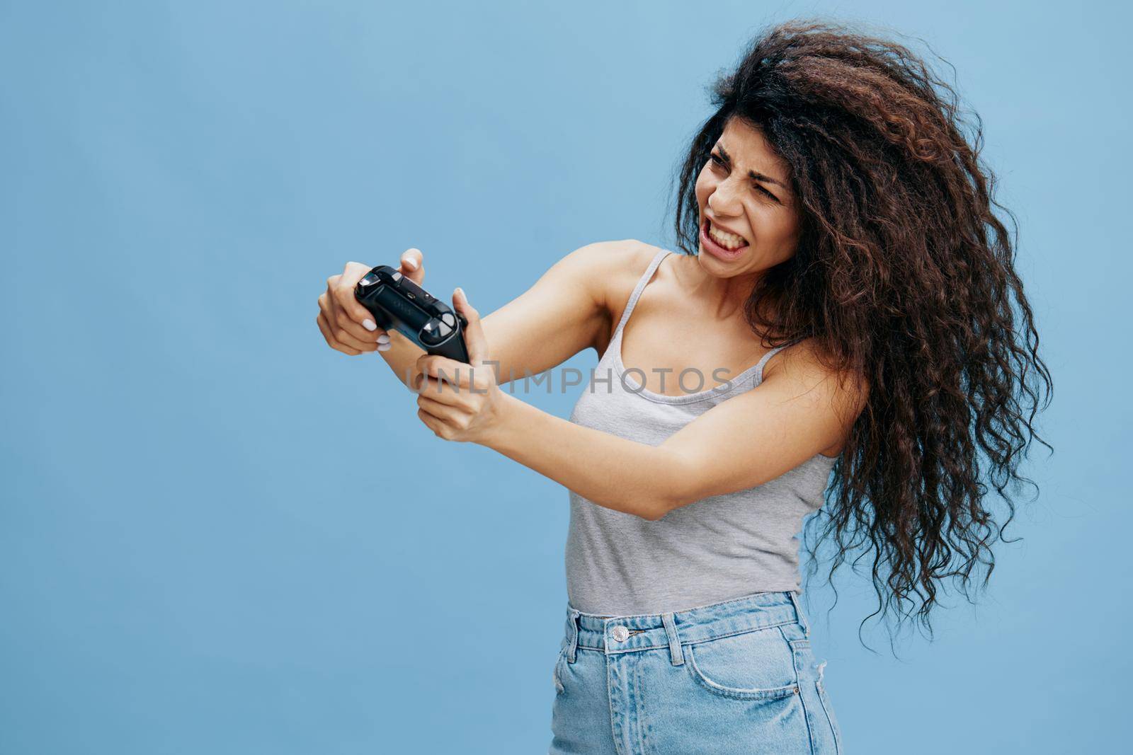 ONLINE GAME AD CONCEPT. Furious beautiful curly Latin lady hold game pad near face, ready to play cool RPG, look at camera. Gaming accessories brands ad. Studio shoot isolated blue background by SHOTPRIME