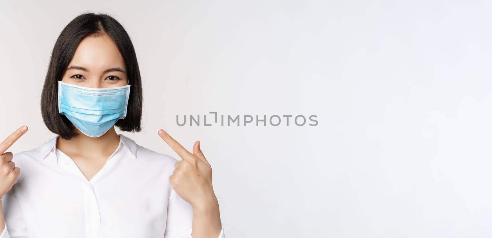 Image of young asian woman pointing at herself while wearing medical face mask, concept of covid-19 protection, standing over white background.