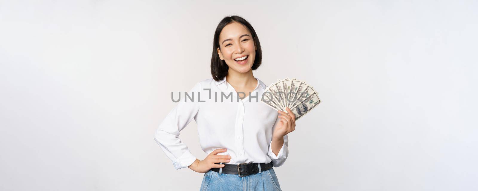 Credit and loan concept. Smiling young asian woman holding cash dollars and looking happy at camera, white background by Benzoix
