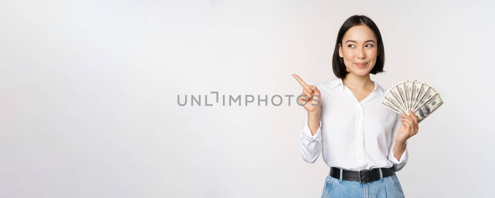 Smiling young modern asian woman, pointing at banner advertisement, holding cash money dollars, standing over white background by Benzoix