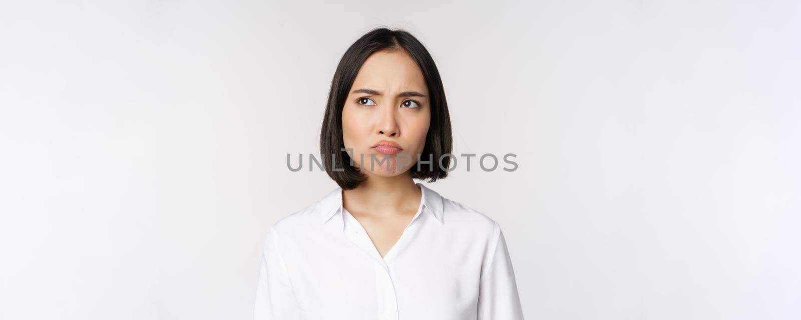 Close up head portrait of young asian woman looking upset and disappointed at left copy space, grimacing and frowning displeased, white background.