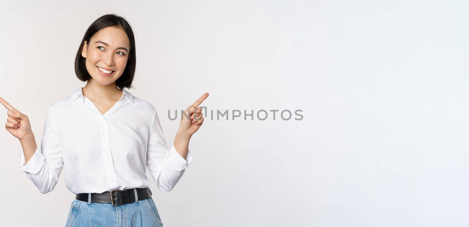 Image of beautiful asian woman pointing fingers left and right, making decision, showing two variants choices, standing over white background.
