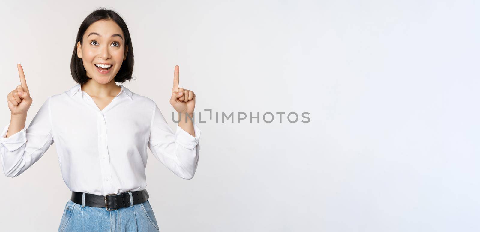 Enthusiastic asian business woman pointing, looking up with happy smiling face, showing company logo or banner, standing over white background by Benzoix