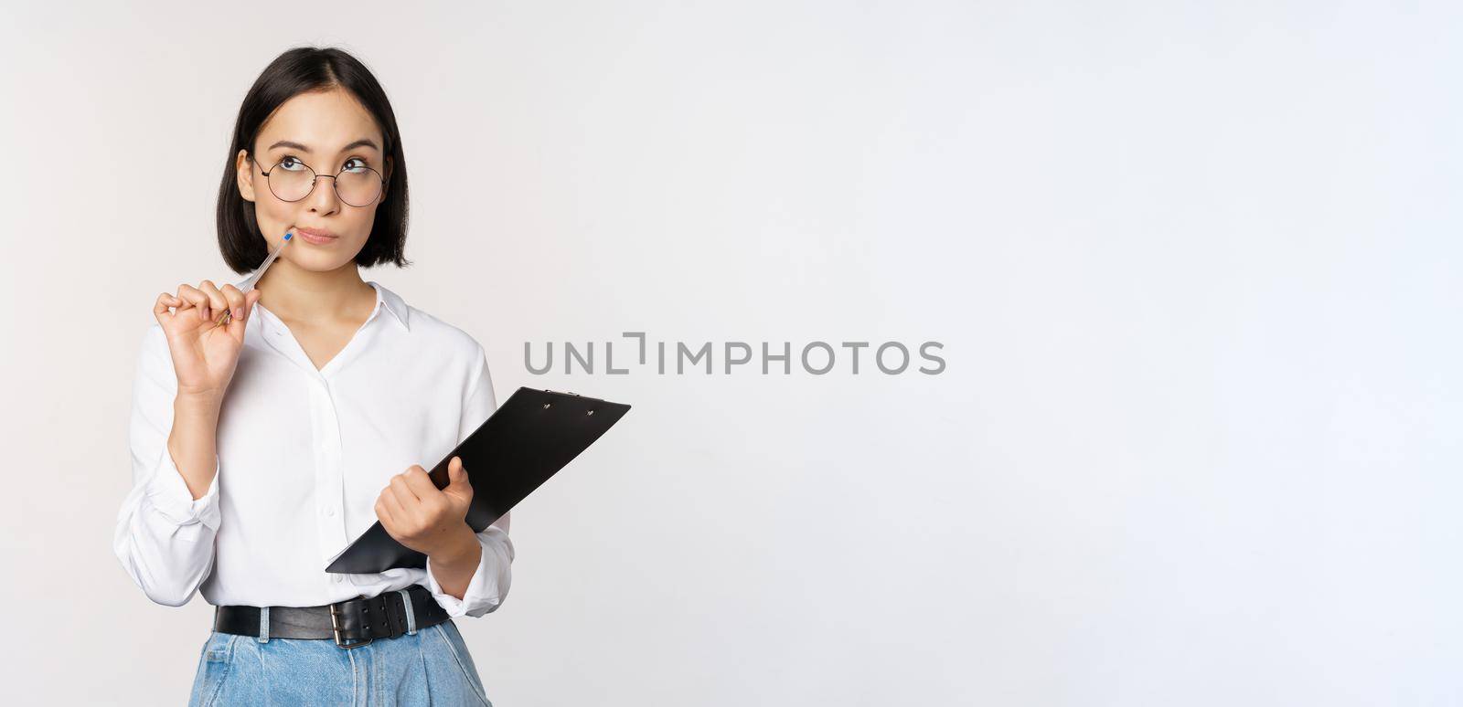 Asian girl in glasses thinks, holds pen and clipboard, writing down, making notes, standing over white background by Benzoix